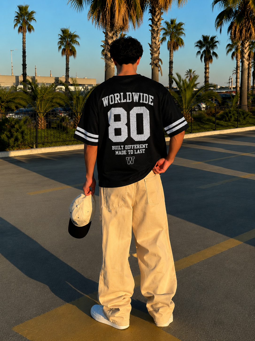 Person wearing a black jersey with 'Worldwide 80' text, standing in a parking lot with palm trees in the background.