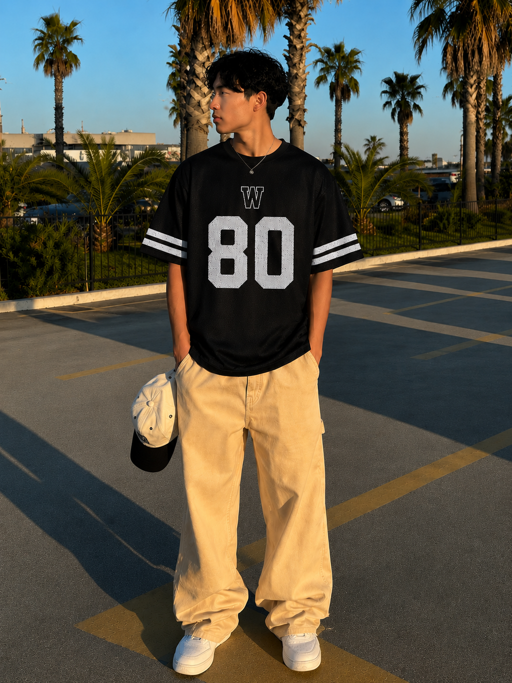 Person wearing a black jersey with white lettering and numbers, standing in a parking lot with palm trees in the background.